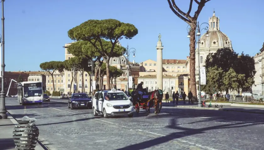 Carrozza trainata da cavalli, taxi e autobus sfilano su un lastricato romano, con la cupola di una chiesa e un monumento antico sullo sfondo, nella zona Battistini Roma.
