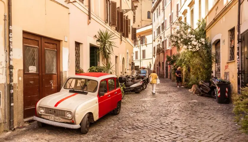Un'auto d'epoca bianca e rossa parcheggiata in un vicolo acciottolato del **quartier Trastevere**. Persone passeggiano, un cane annusa l'aria.