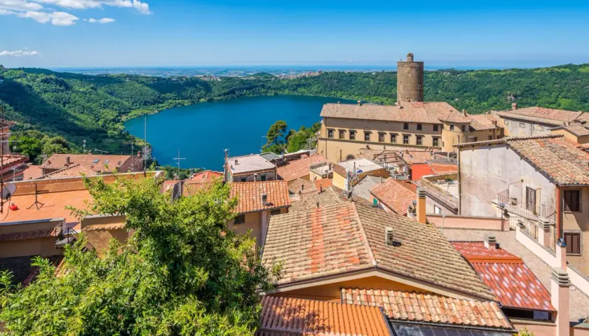 Panoramica dei colli di Roma oggi: un lago azzurro circondato da colline verdi e un borgo con tetti rossi e una torre medievale.