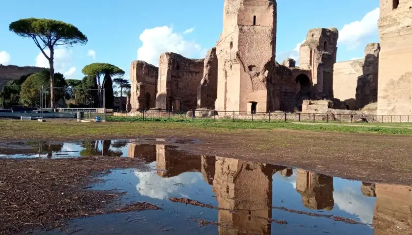 Le maestose rovine delle **Terme di Caracalla** si specchiano in una pozza d'acqua, sotto un cielo azzurro con nuvole bianche.