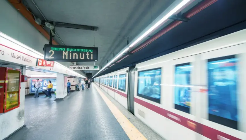 Stazione metro Roma: un treno in arrivo tra 2 minuti. Persone attendono sul binario, un cartello indica "uscita/exit".