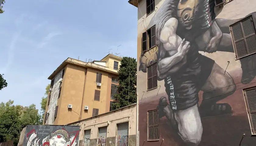 Street art a Tor Marancia: un imponente murale raffigura un lottatore con maschera tribale, mentre su un muro più basso un'altra opera celebra la cultura orientale.
