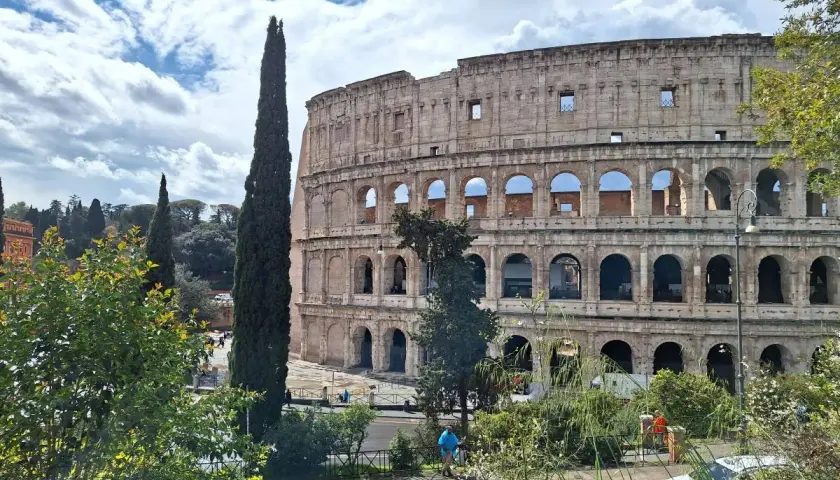 Le maestose **colonne di Roma** del Colosseo si stagliano contro un cielo azzurro con nuvole bianche. Alberi verdi e un'auto bianca completano la scena.