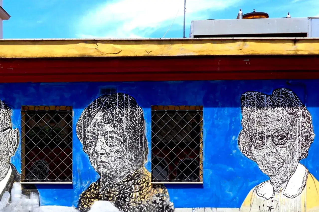 Volti stilizzati in bianco e nero su un muro blu acceso, parte di un **murales ostiense**. Le finestre con grate interrompono la composizione.