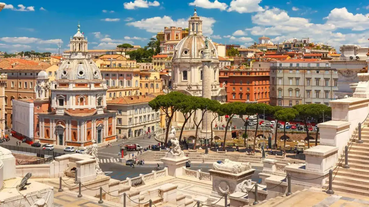 Vista panoramica di Roma con l'Altare della Patria in primo piano, cupole imponenti e palazzi storici sotto un cielo azzurro con nuvole bianche.