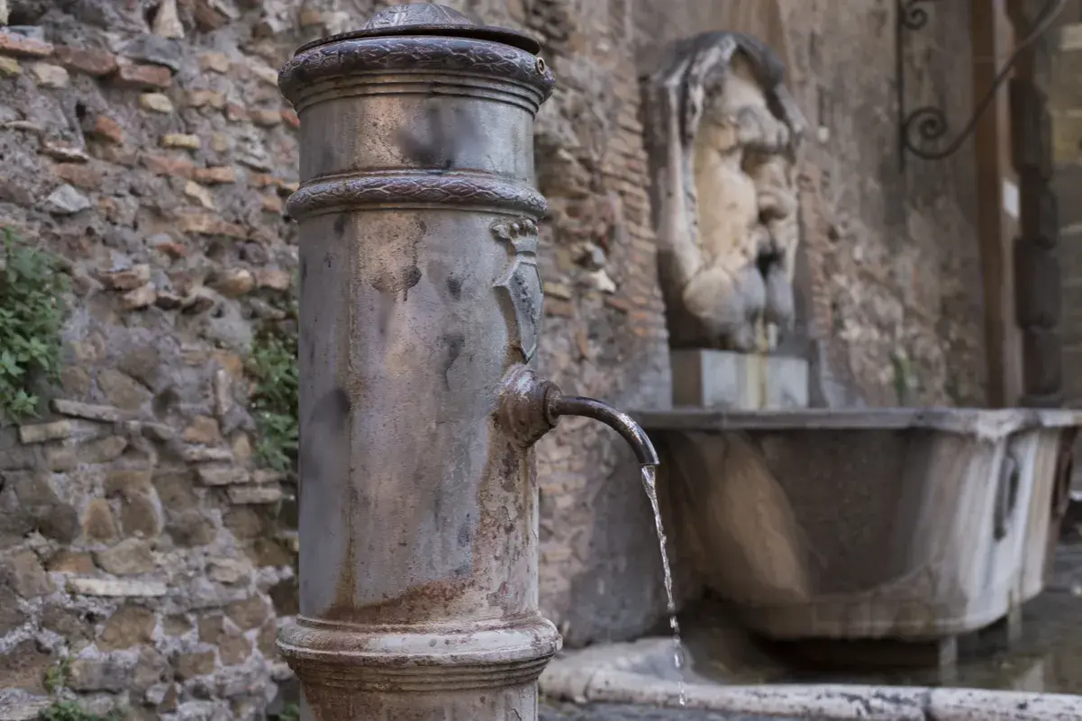Fontanella romana con un nasone che eroga acqua fresca. Sullo sfondo, un antico volto scolpito.