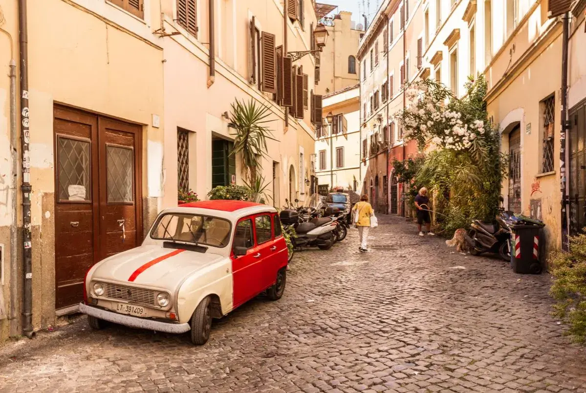 Un'auto d'epoca bianca e rossa parcheggiata in un vicolo acciottolato del **quartier Trastevere**. Persone passeggiano, un cane annusa l'aria.