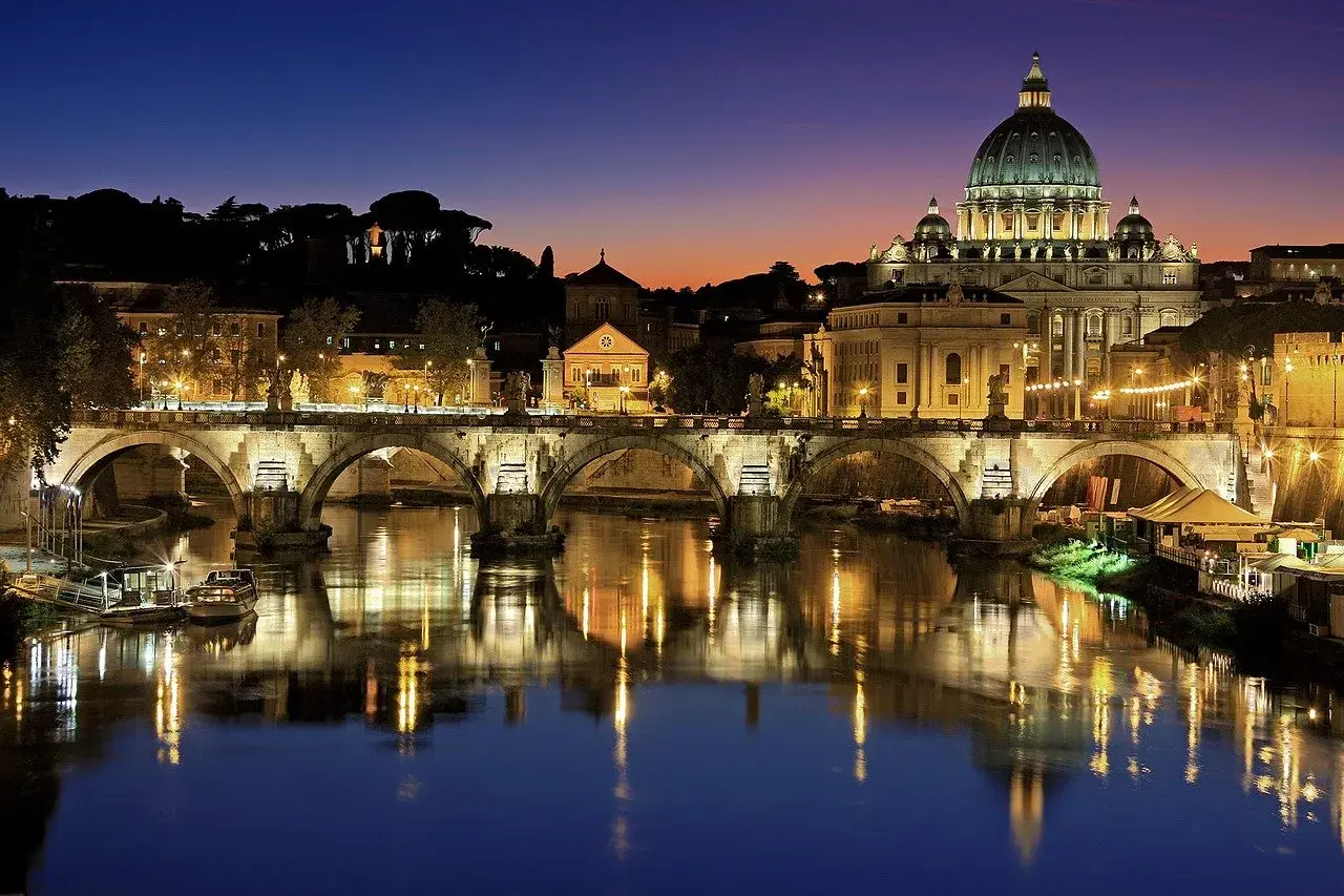 Ponte Sant'Angelo illuminato di notte, con la cupola di San Pietro sullo sfondo. Un'idea per cosa visitare a Roma in due giorni.