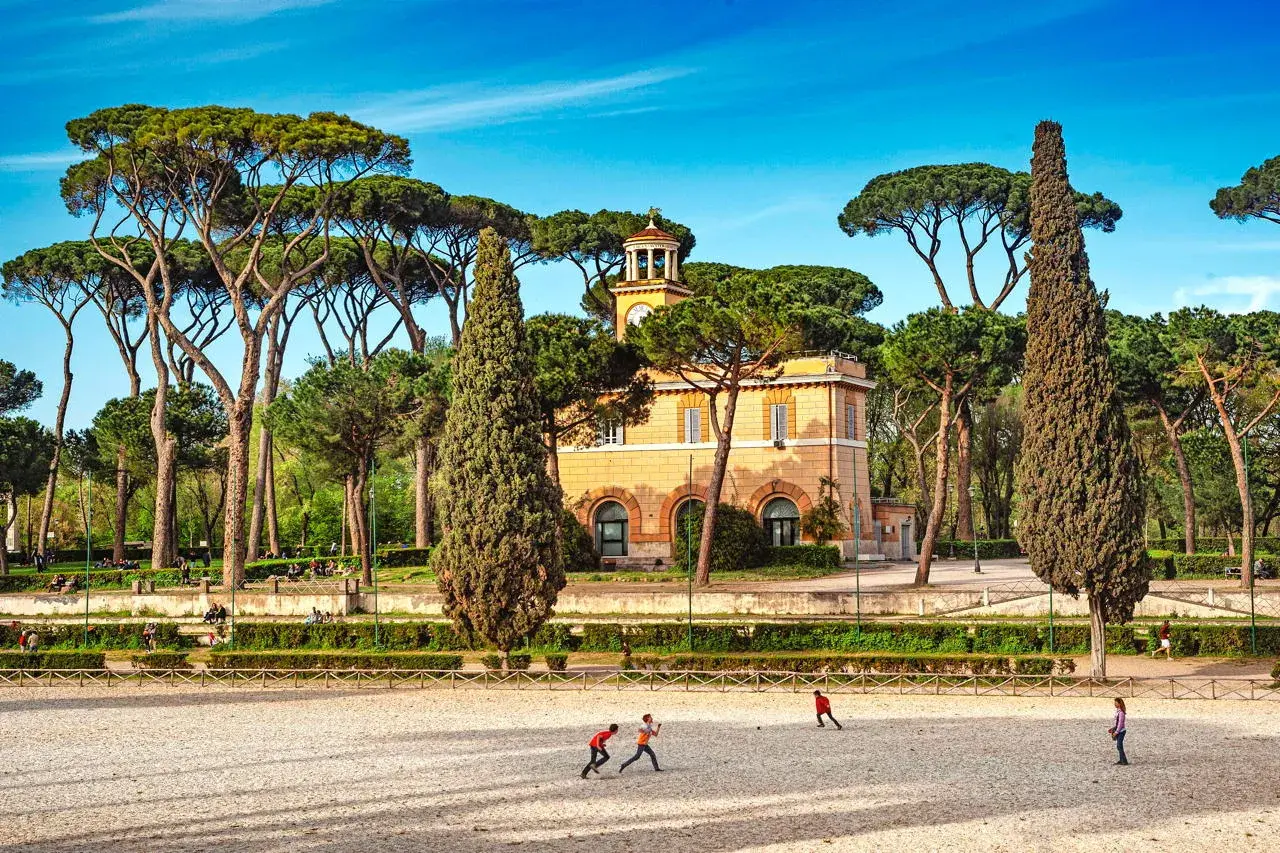Un edificio giallo con una torre dell'orologio, circondato da pini marittimi e cipressi. Bambini giocano in un'area sabbiosa. Ecco cosa vedere ai Parioli.