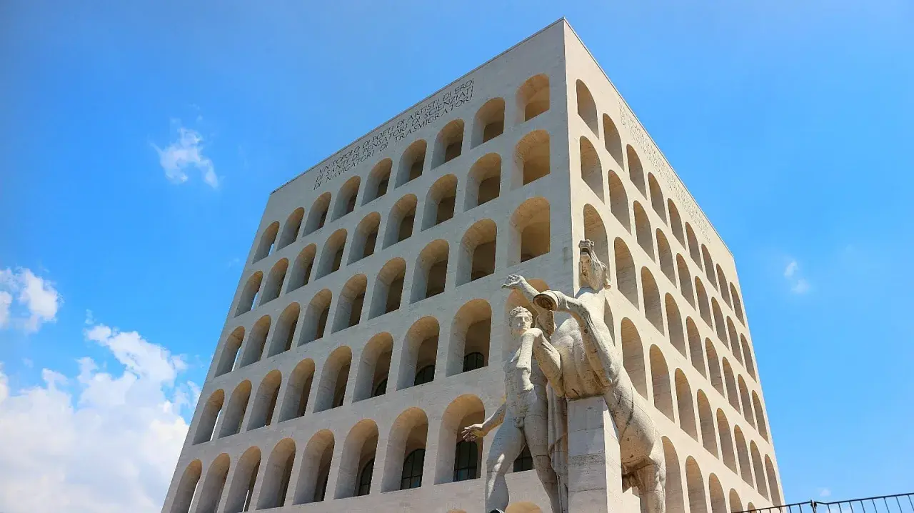 Palazzo Mussolini, con la sua facciata ad archi sovrapposti, domina il cielo azzurro. Una scultura di uomini e cavalli si erge in primo piano.