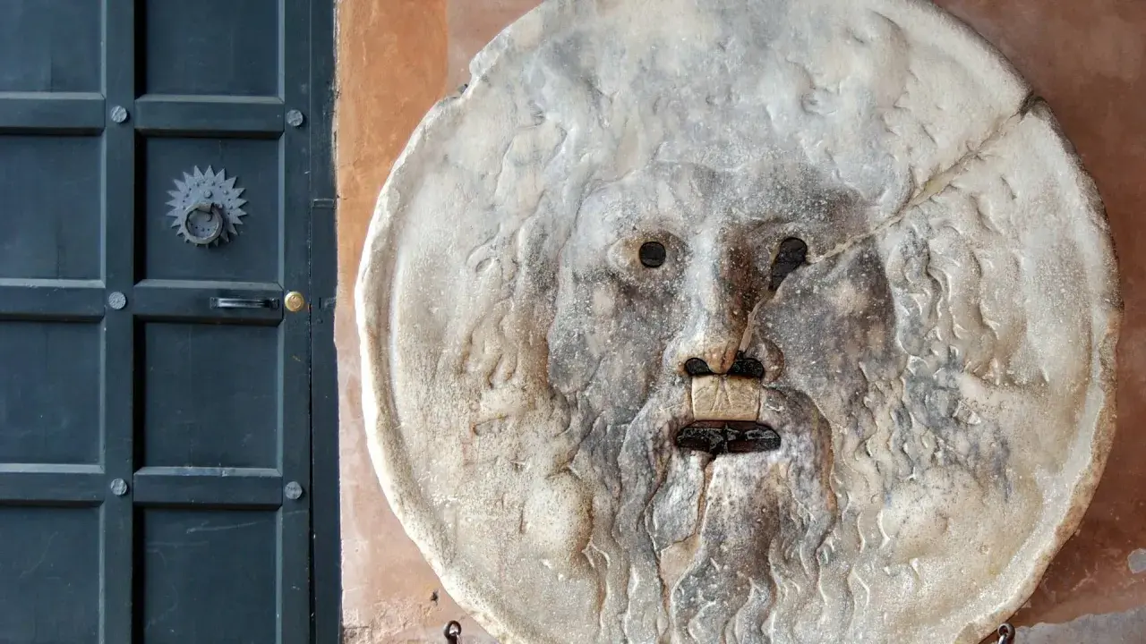 La Bocca della Verità, un volto scolpito nella pietra, sembra pronto a giudicare chiunque osi mentire. Accanto, una porta scura.