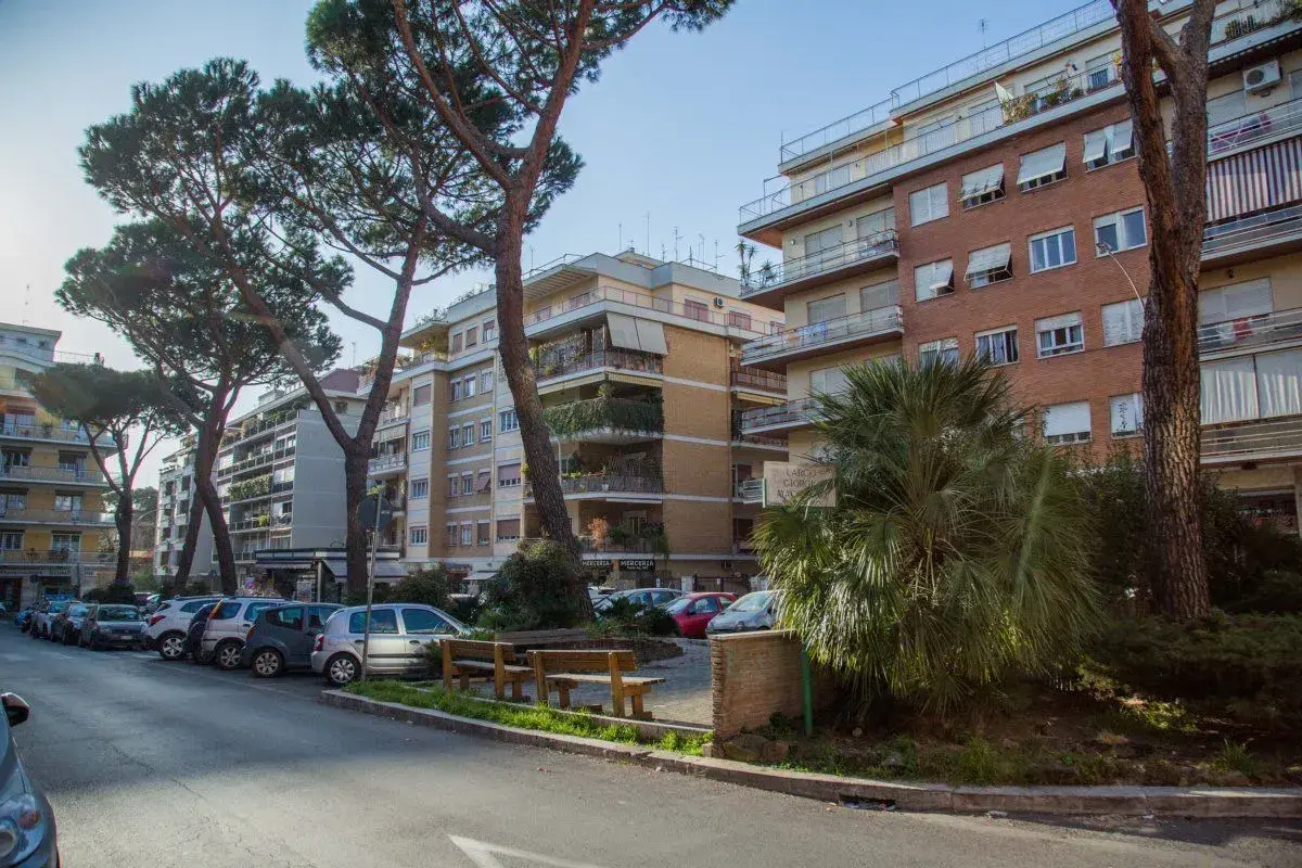 Edifici residenziali e pini marittimi nel quartiere Balduina a Roma. Auto parcheggiate lungo la strada, panchine e vegetazione rigogliosa.