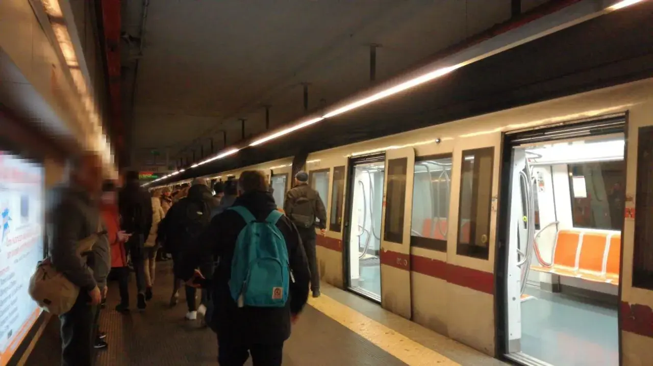 Persone in attesa sul binario della metro domenica, con un treno bianco e rosso fermo con le porte aperte.