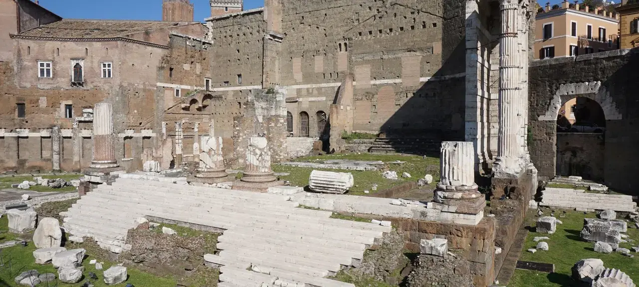 Rovine del Foro di Augusto a Roma, con imponenti colonne e scalinate di marmo bianco che emergono dall'erba verde.