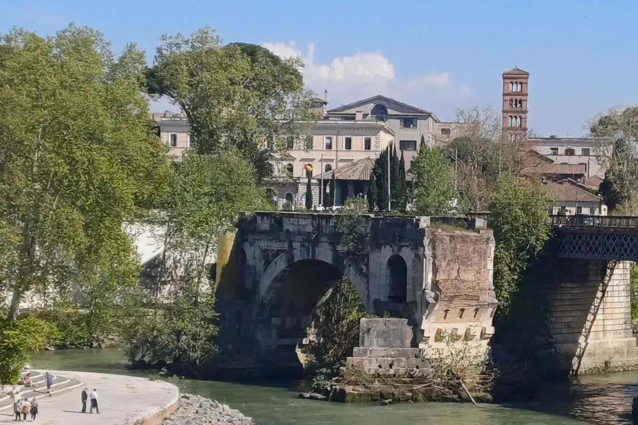 Antico **ponte rotto a Roma**, con rovine e vegetazione che si arrampicano sui resti. In lontananza, edifici storici e una torre campanaria.