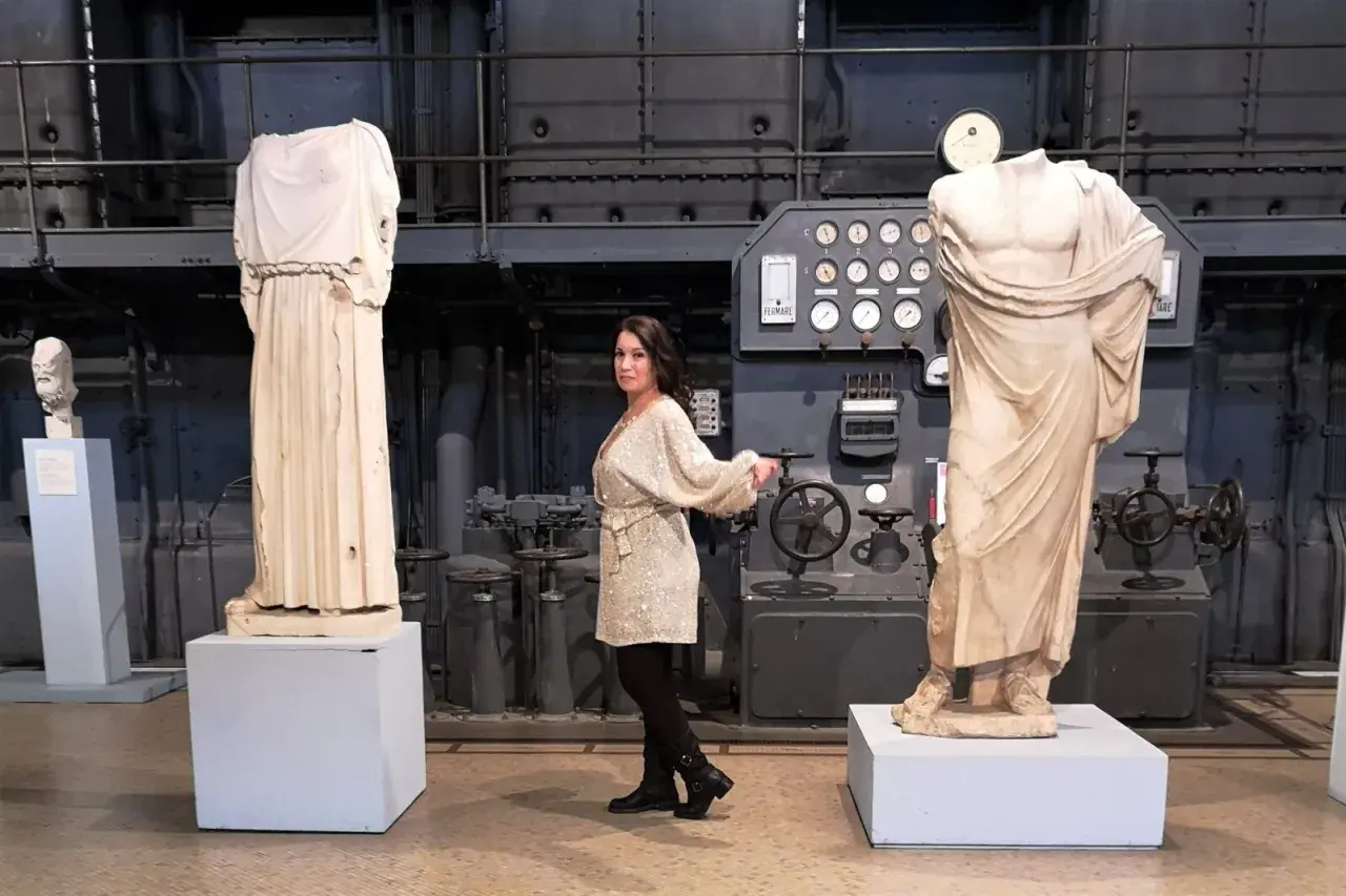 Donna in abito scintillante tra statue classiche e macchinari industriali, forse nel vecchio gasometro.