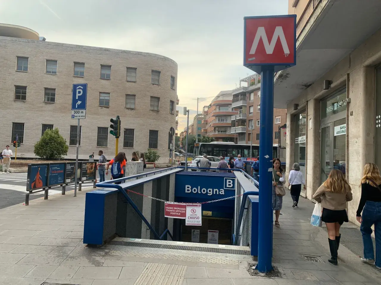 Ingresso della metro Bologna a Roma, con insegna rossa e bianca. Persone camminano sul marciapiede.