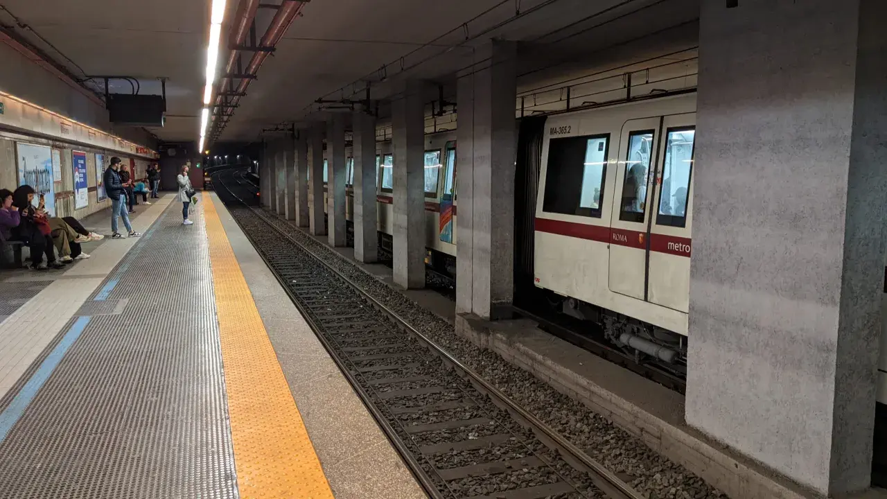 Persone attendono il treno alla stazione metro a Lepanto. Un convoglio bianco e rosso è fermo sui binari.