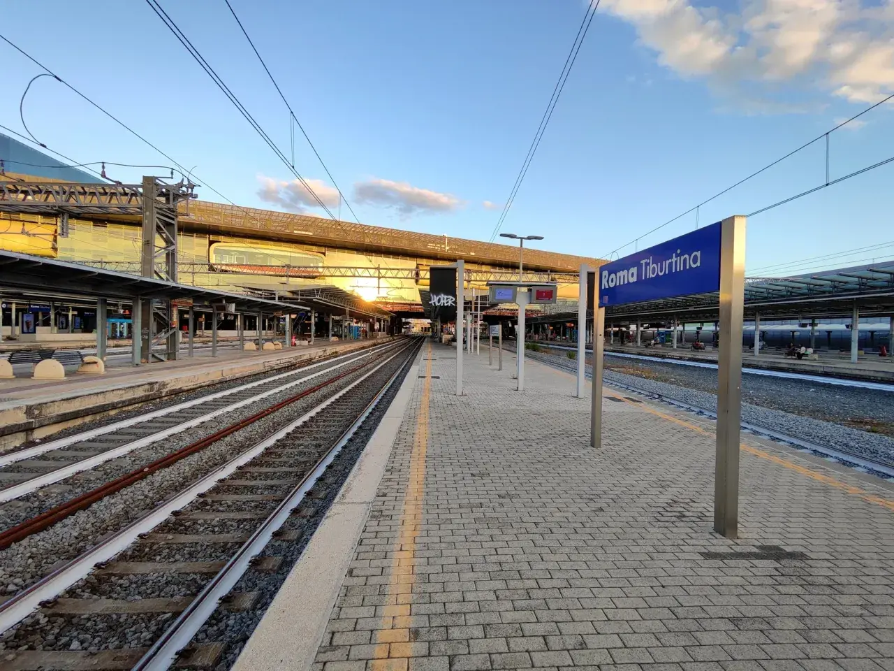 Binari e pensiline alla stazione Roma Tiburtina, con il sole che illumina la moderna architettura.