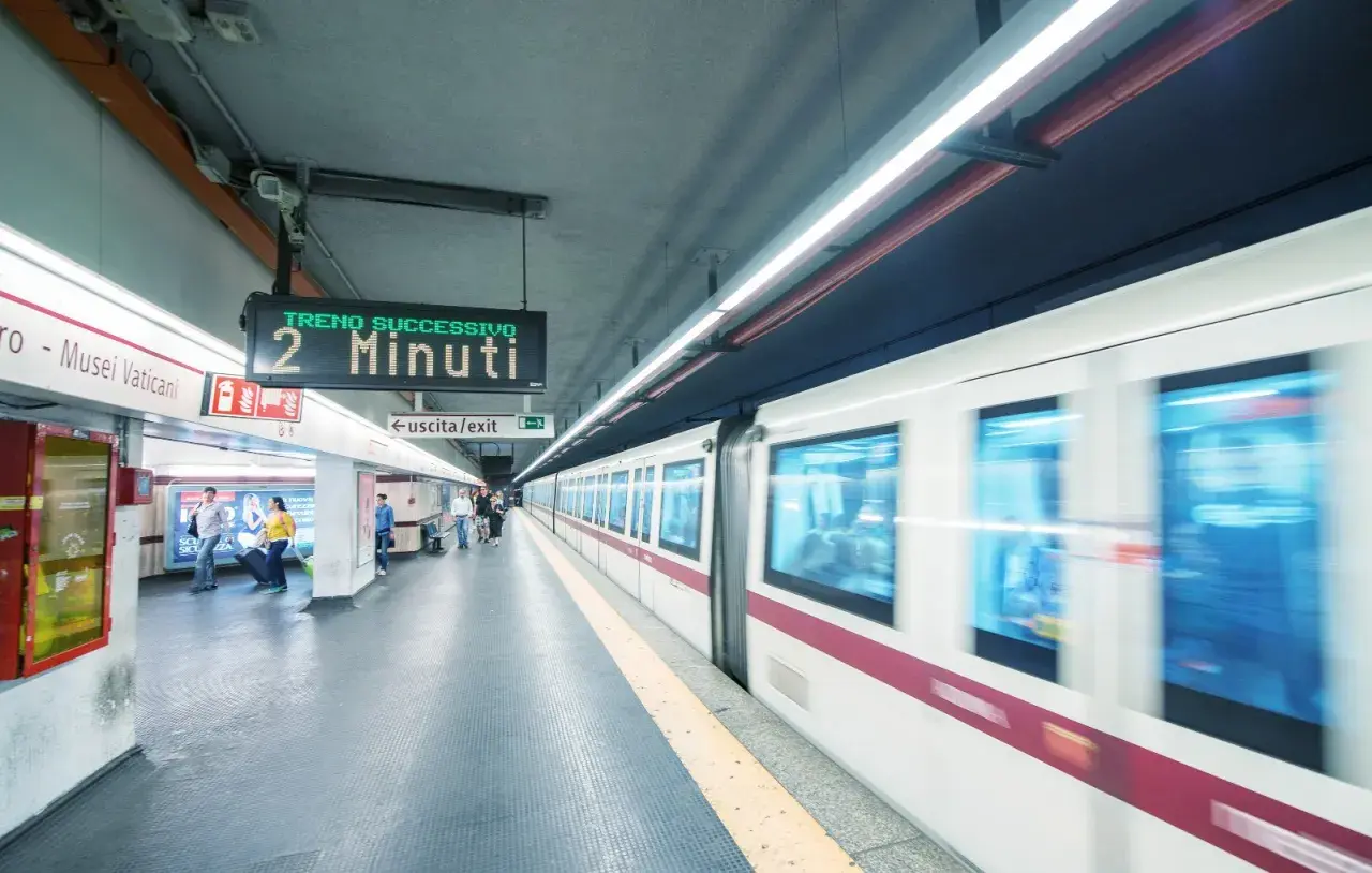 Stazione metro Roma: un treno in arrivo tra 2 minuti. Persone attendono sul binario, un cartello indica "uscita/exit".