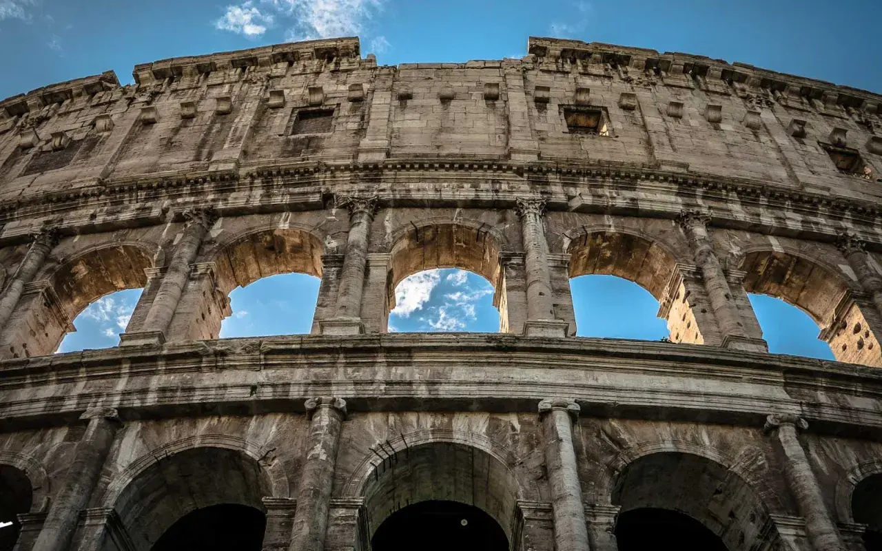 Il Colosseo visto dal basso, con il cielo azzurro e nuvole tra gli archi. Un'ottima meta per chi cerca come arrivare al Colosseo da Stazione Termini.