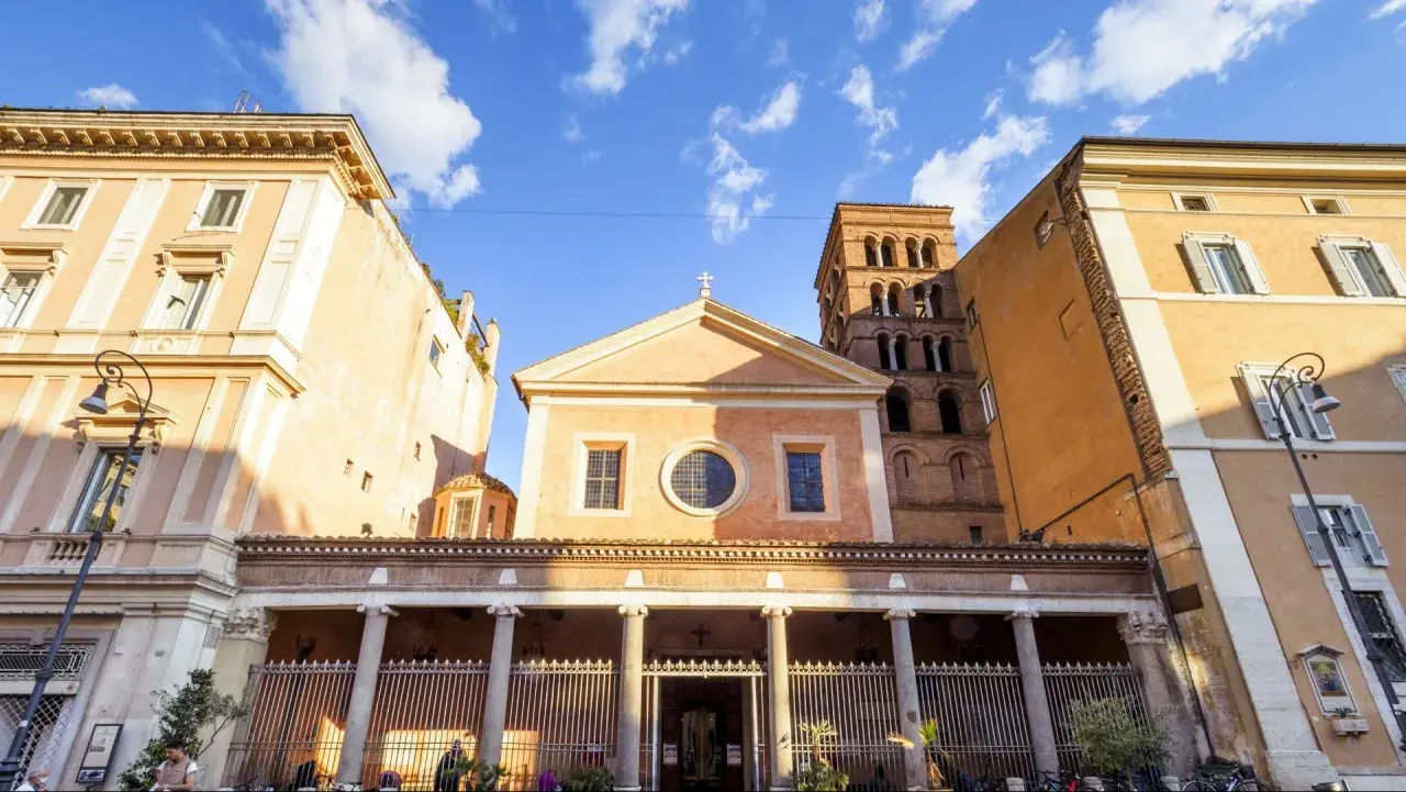 Facciata di San Lorenzo in Lucina a Roma, con il suo campanile romanico e palazzi circostanti sotto un cielo azzurro con nuvole bianche.