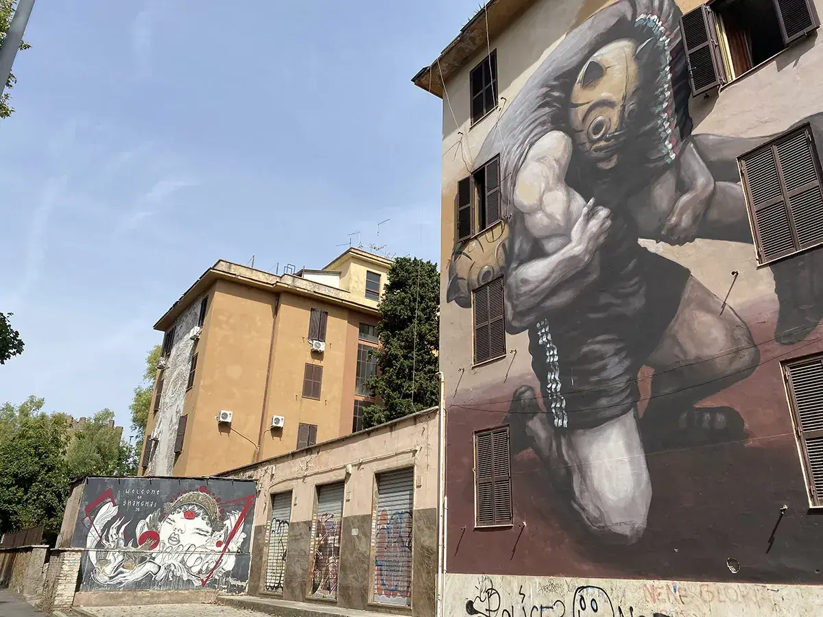Street art a Tor Marancia: un imponente murale raffigura un lottatore con maschera tribale, mentre su un muro più basso un'altra opera celebra la cultura orientale.