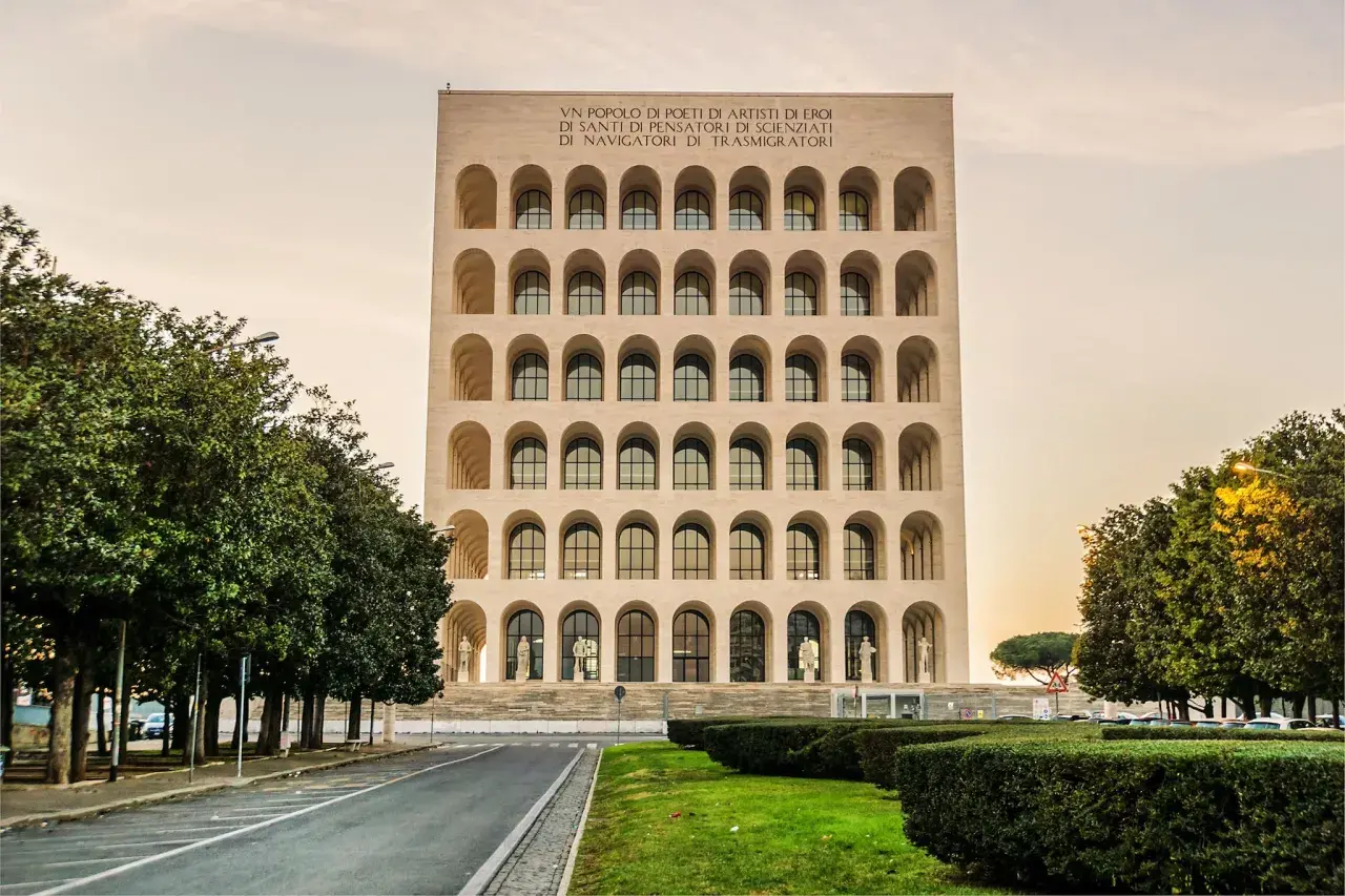 Edificio con arcate sovrapposte, simile a una stazione metro EUR Magliana, con iscrizione in latino e alberi verdi.