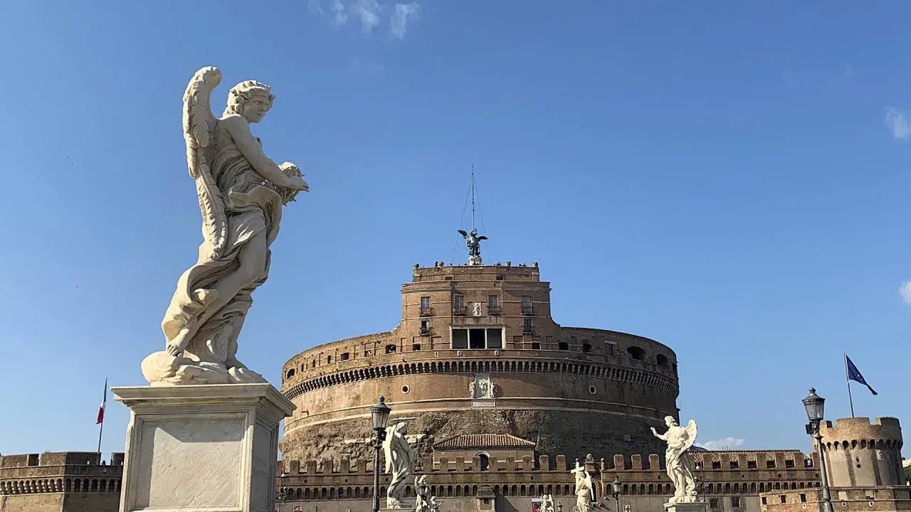 Un angelo scolpito domina la scena davanti al maestoso mausoleo di Roma, Castel Sant'Angelo, sotto un cielo azzurro.