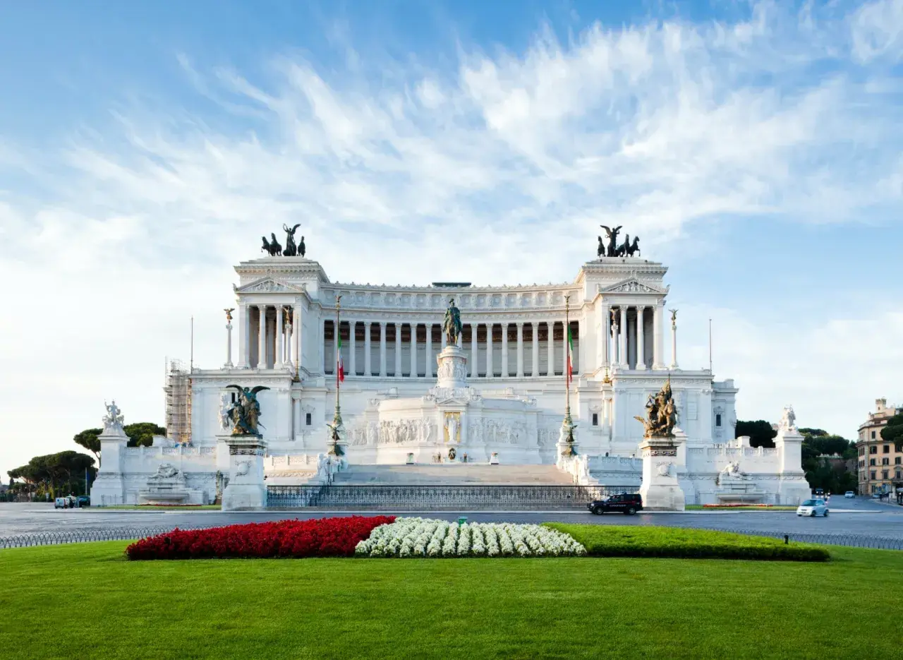 Il Vittoriano, monumento imponente, si erge maestoso. Vicino alla fermata metro Altare della Patria, un prato verde e fiori rossi e bianchi creano un contrasto vibrante.