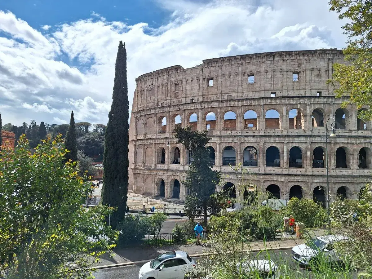 Le maestose **colonne di Roma** del Colosseo si stagliano contro un cielo azzurro con nuvole bianche. Alberi verdi e un'auto bianca completano la scena.