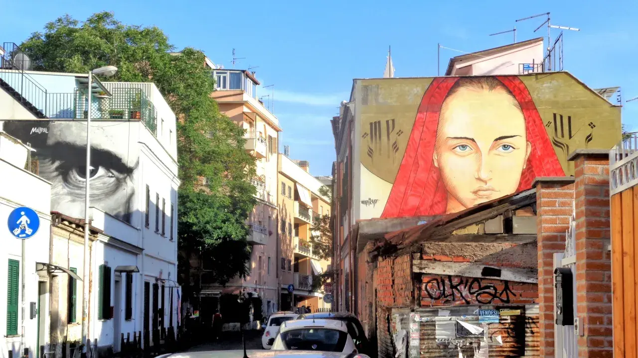 Un grande volto femminile con un velo rosso domina un edificio nel quartiere Pigneto. Accanto, un occhio gigante osserva la strada.