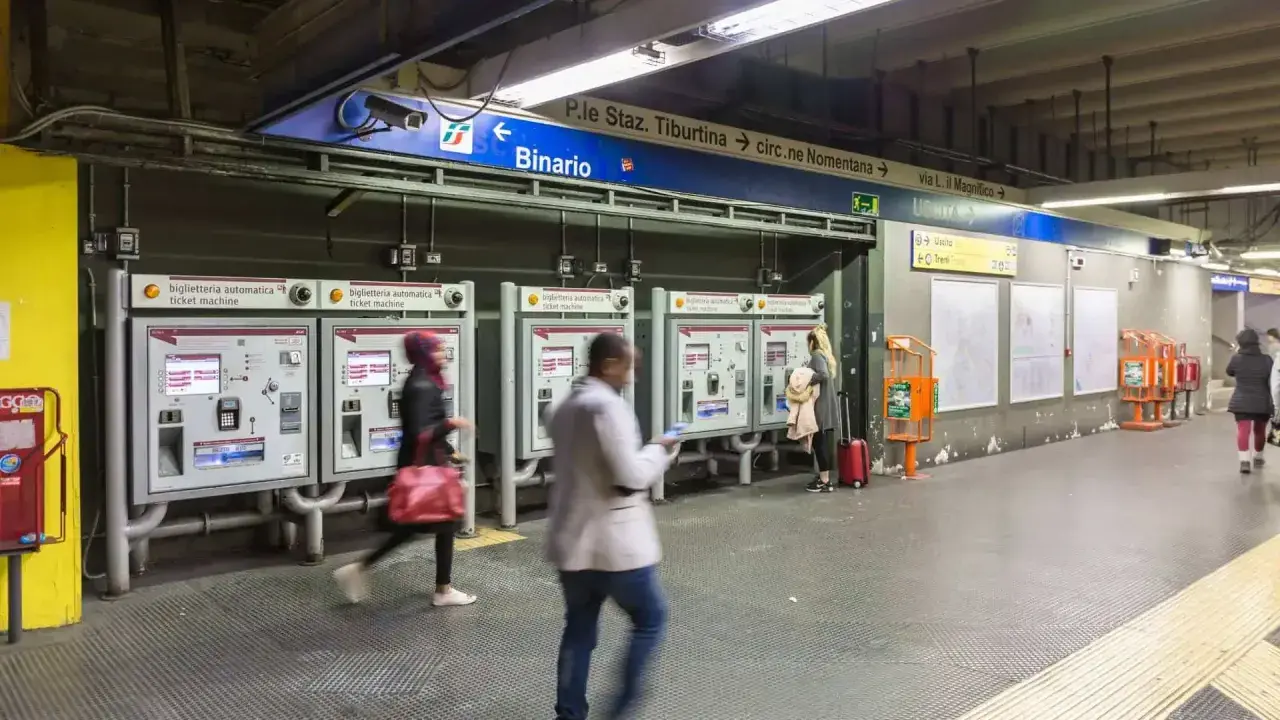 Persone acquistano biglietti alle macchinette automatiche nella stazione metro Roma EUR. Segnali indicano binari e uscite.