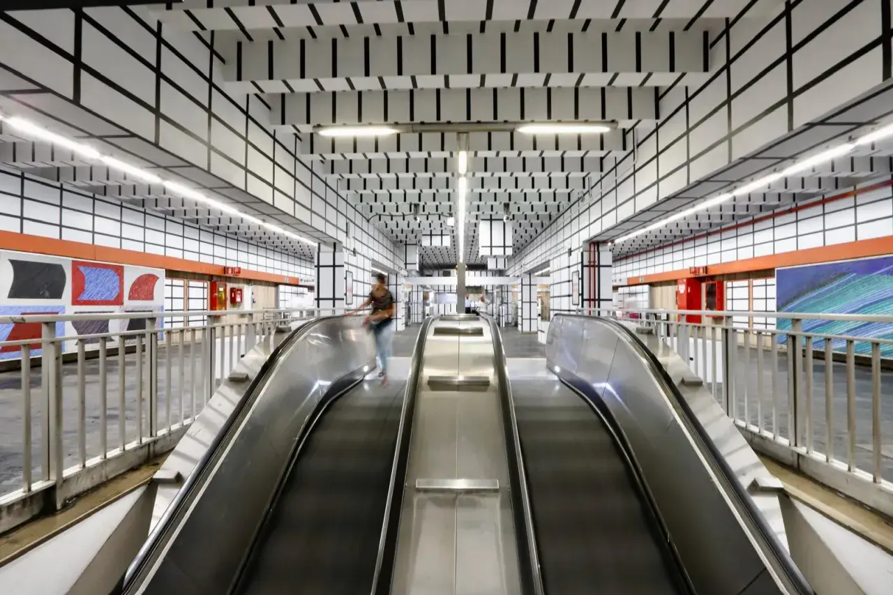 Scale mobili nella moderna stazione metro Vittorio Emanuele a Roma, con soffitto a griglia nera su bianco e opere d'arte colorate alle pareti.