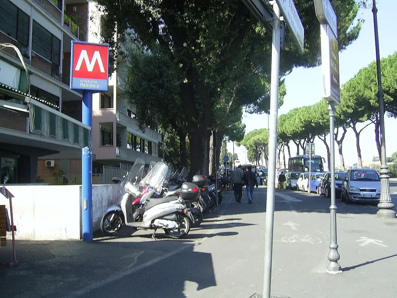 Segnale della metro Rebibbia a Roma, con scooter parcheggiati e alberi lungo la strada.