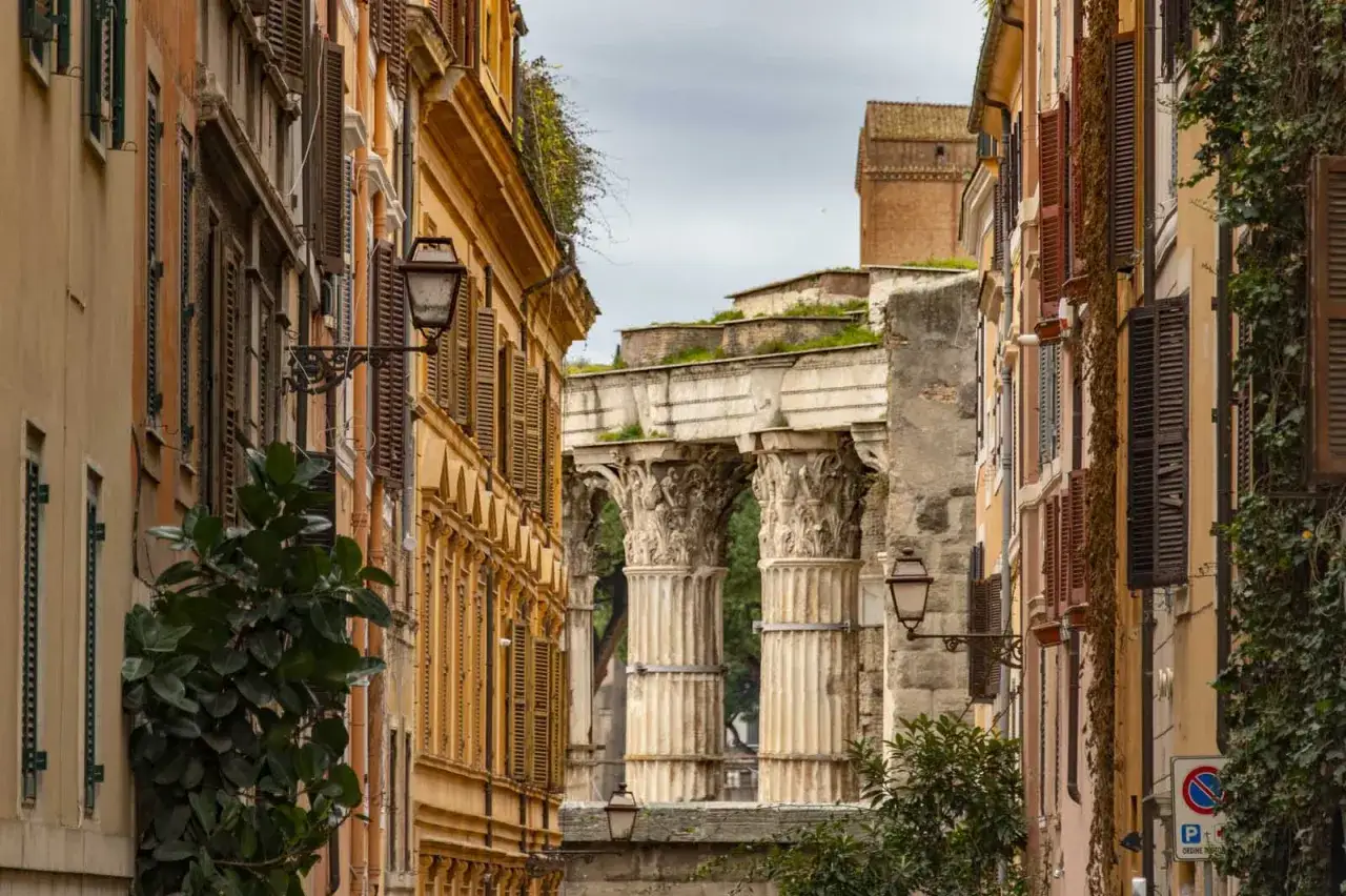 Antiche rovine romane, forse la Suburra, tra palazzi color ocra con persiane chiuse e lampioni.