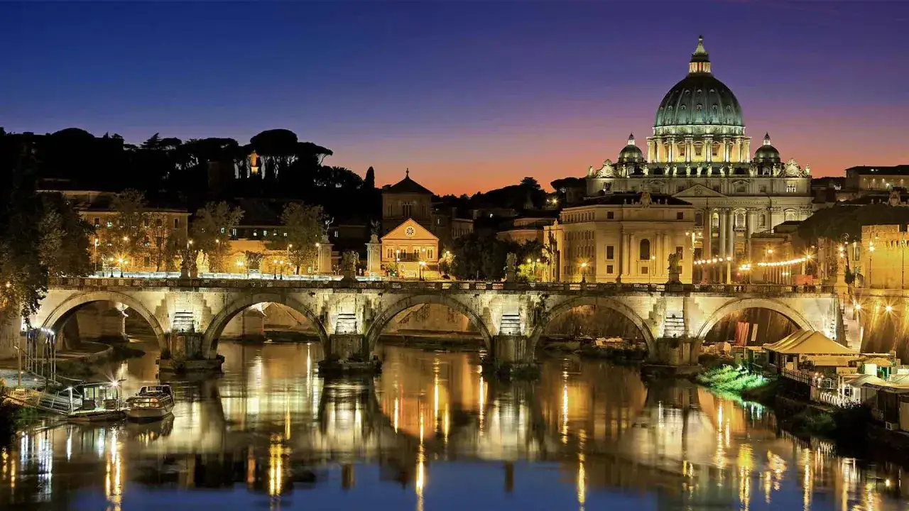 Ponte Sant'Angelo illuminato di notte, con la cupola di San Pietro e Paolo sullo sfondo. Riflessi sull'acqua del Tevere.