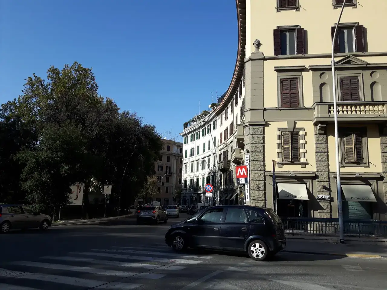 Strada trafficata in un quartiere di Roma, con edifici storici e un'insegna della metropolitana.