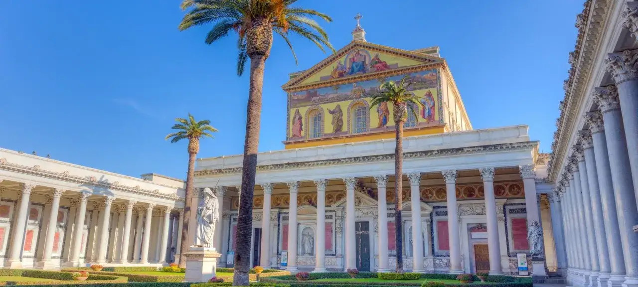 La Basilica Papale di San Paolo fuori le Mura, con le sue maestose colonne e palme, si erge sotto un cielo azzurro.