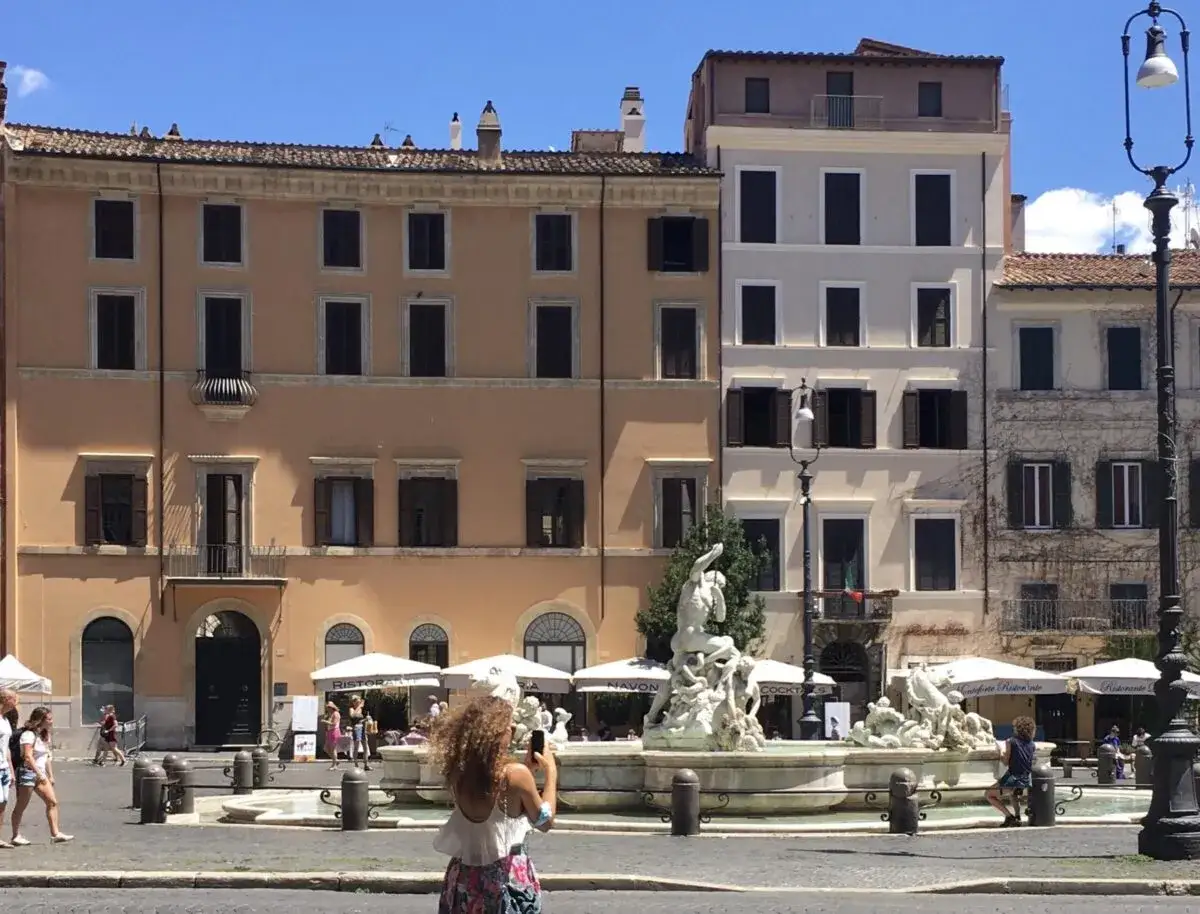 Un palazzo storico domina Piazza Navona, con una fontana barocca al centro. Una turista scatta una foto.