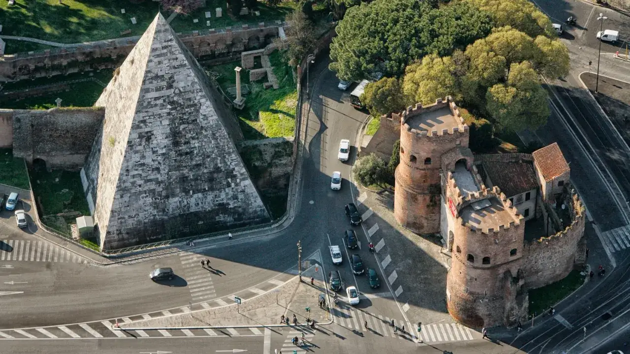 Piramide Cestia e Porta San Paolo nel quartiere Ostiense, Roma, con auto e pedoni che attraversano la strada.