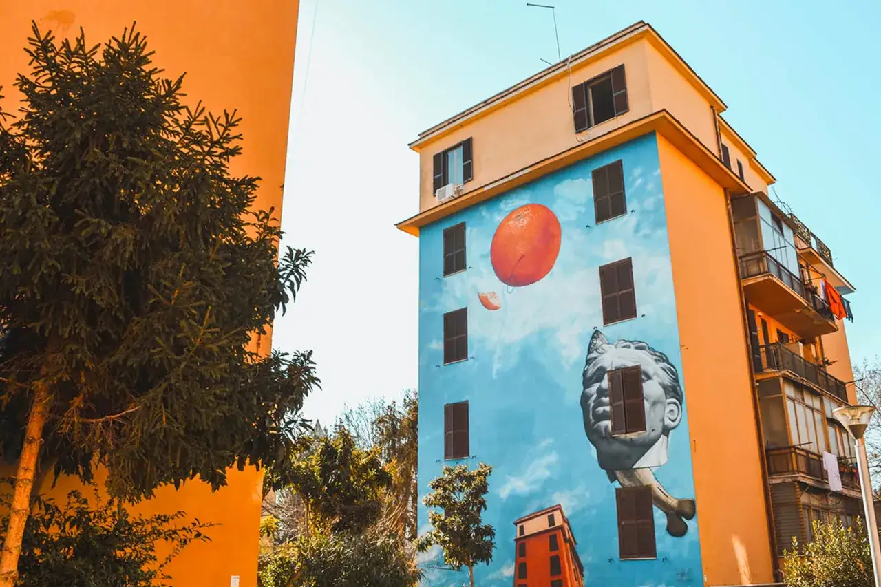 Un palazzo colorato a Tor Marancia con un murale surreale: una testa classica con finestre al posto degli occhi, un palloncino arancione e un cielo azzurro.