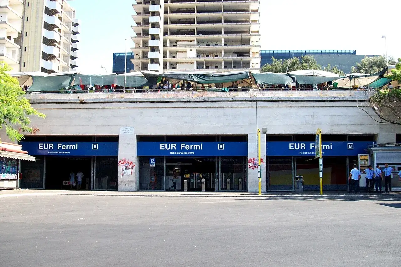 Ingresso della stazione EUR Fermi, con edifici incompiuti sullo sfondo e bancarelle improvvisate sul tetto.