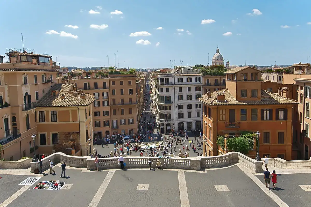 Vista dall'alto di Via del Corso a Roma, con edifici storici, una piazza affollata e la cupola di una chiesa in lontananza.