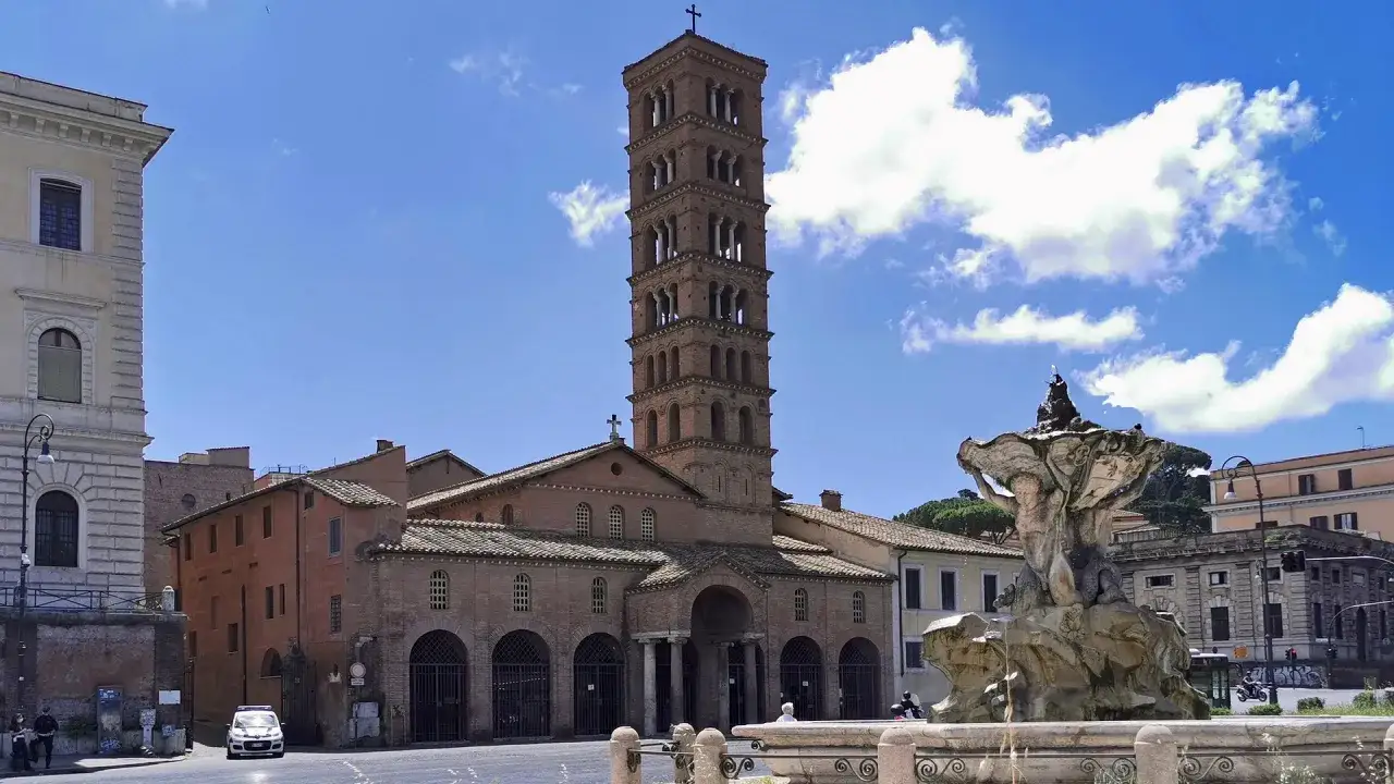 Fontana con la Boca de la Verita e la Basilica di Santa Maria in Cosmedin a Roma, sotto un cielo azzurro con nuvole bianche.