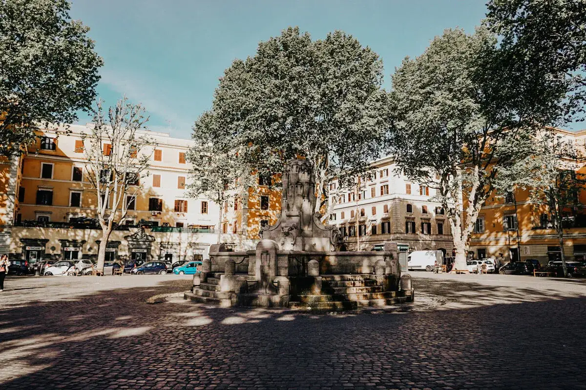 Fontana di Testaccio, Roma, con palazzi gialli e alberi rigogliosi che proiettano ombre sulla pavimentazione in sampietrini.