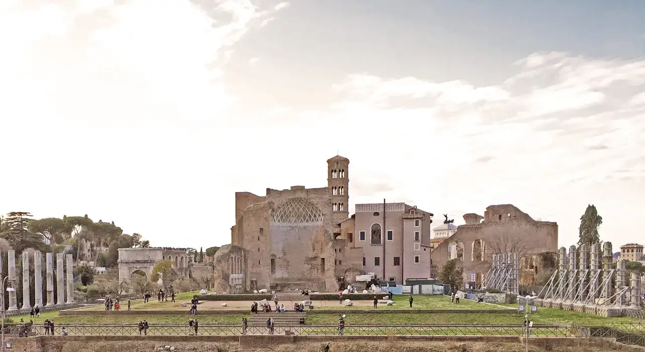 Resti del Foro Romano, con il tempio di Venere Roma in lontananza, sotto un cielo nuvoloso. Turisti passeggiano tra le rovine.