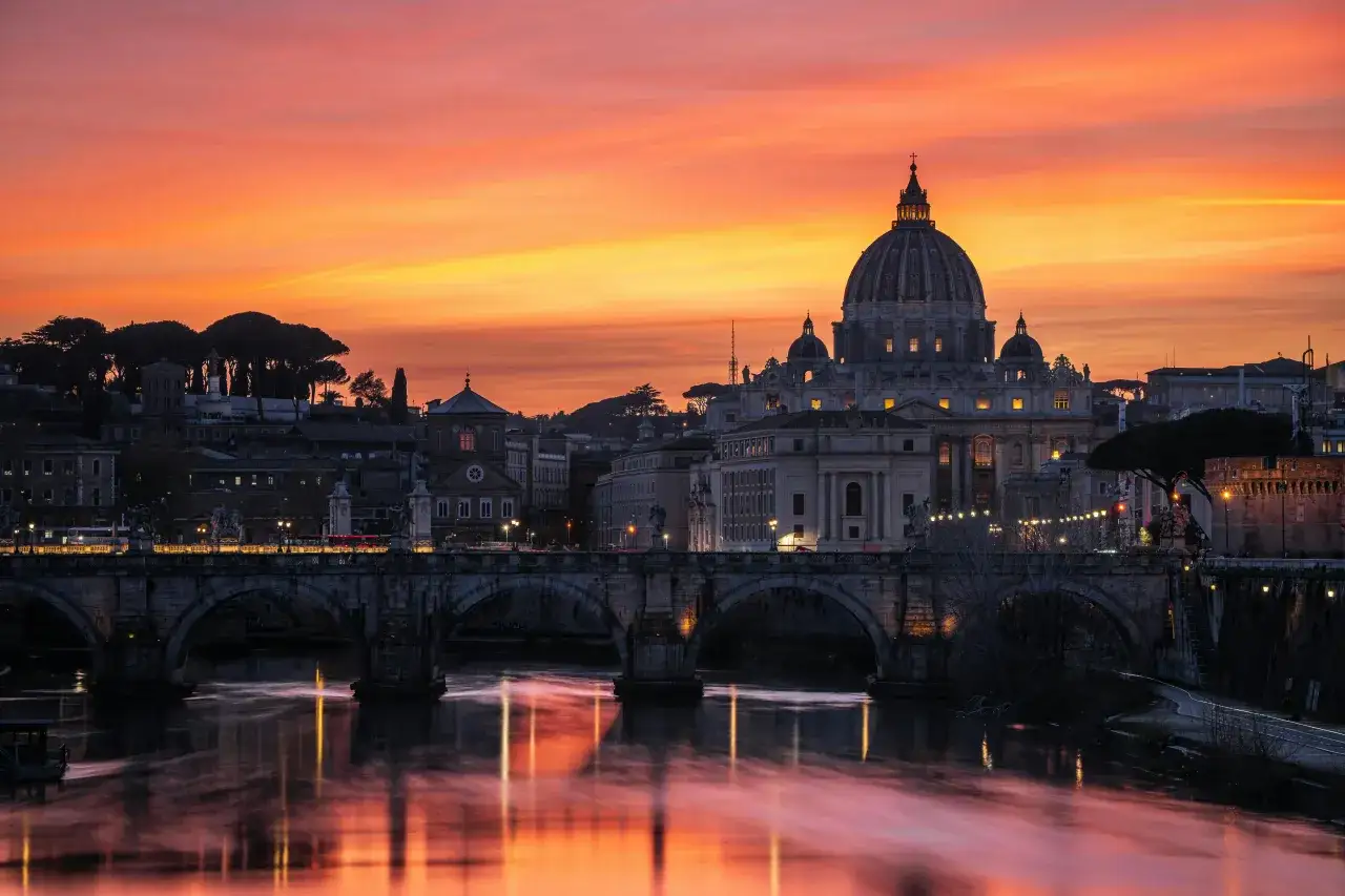 Tramonto infuocato su Roma, con la cupola di San Pietro che domina il cielo. Il Tevere riflette i colori vibranti, un'immagine iconica dell'alta stagione a Roma.