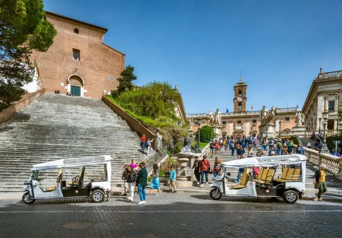 Roma in alta stagione: turisti salgono la scalinata verso il Campidoglio, con golf cart pronti per esplorare la citt&agrave;.