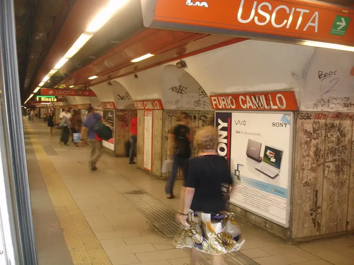 Persone in attesa alla fermata Furio Camillo della metropolitana, con cartelloni pubblicitari e segnali di uscita.